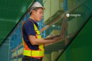A warehouse worker wearing a hard hat and safety vest while using a laptop in front of storage shelves.